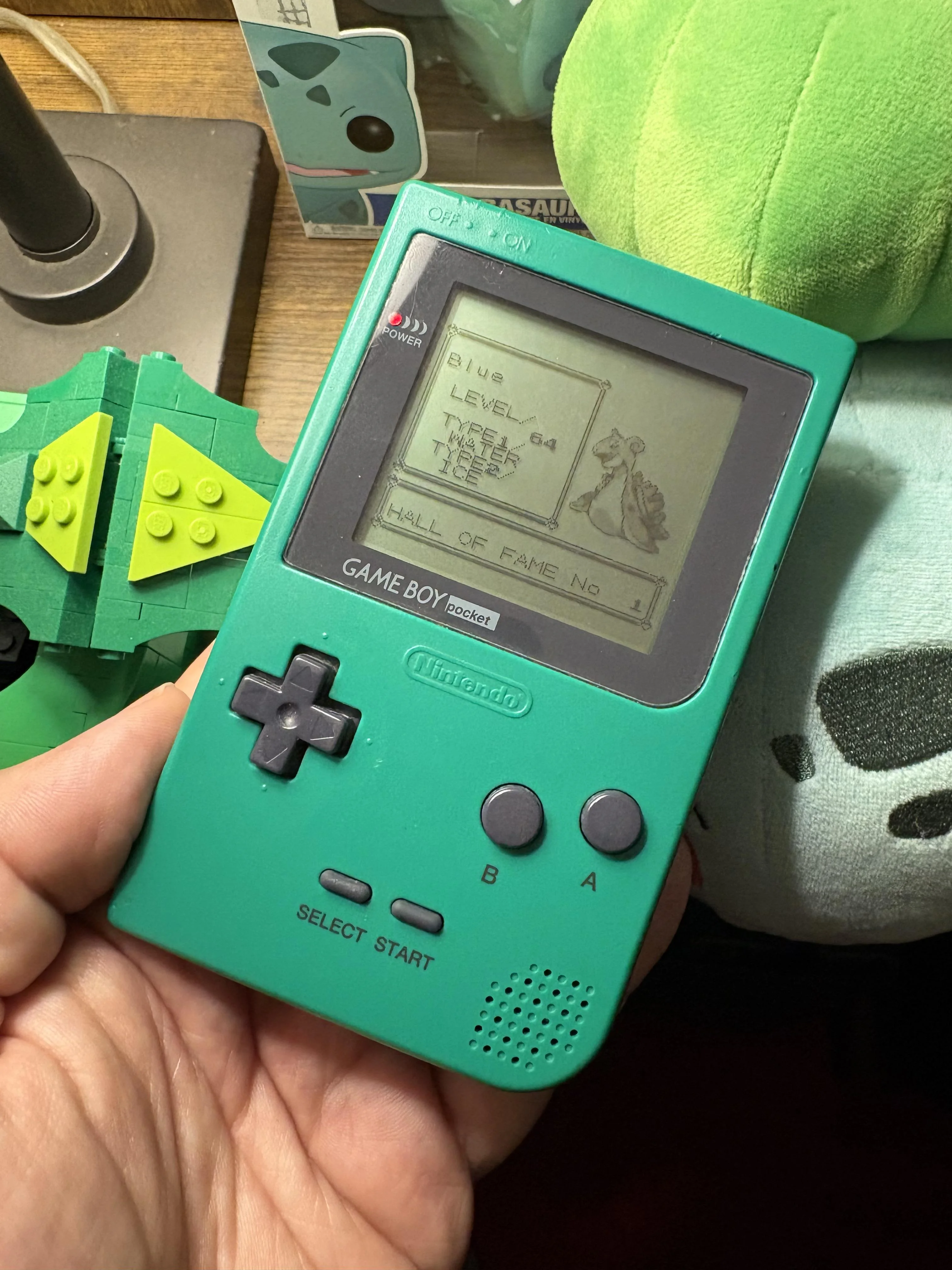 A hand holding a green Game Boy Pocket displaying Pokémon